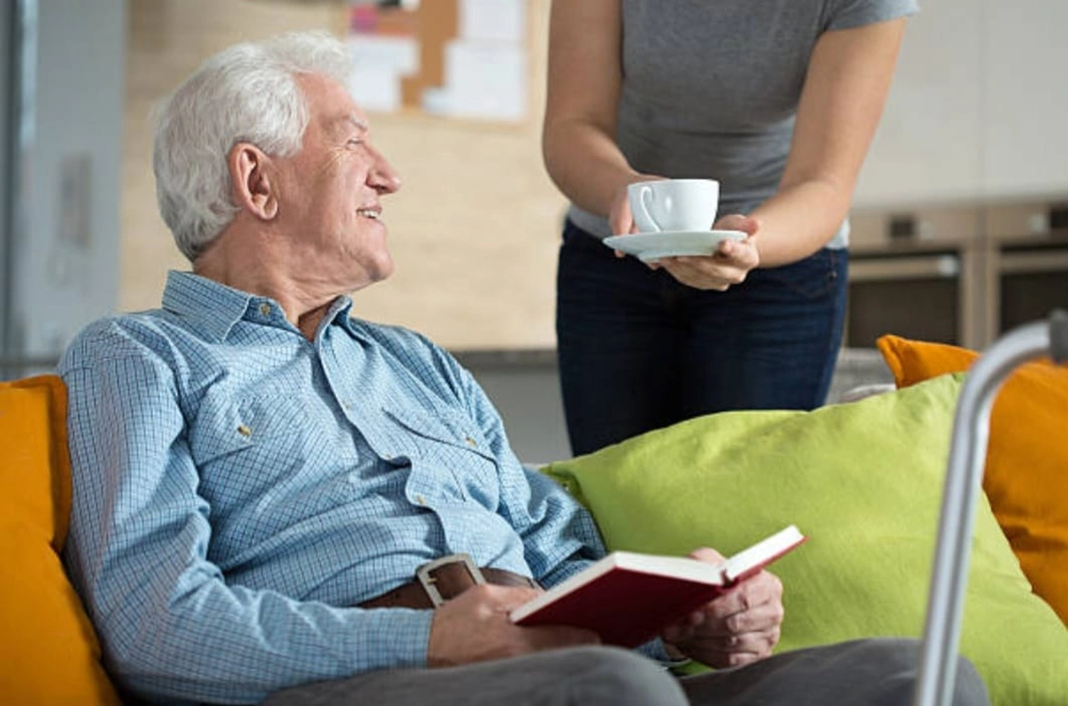 Mbaye Jeng – Companionship Service - Dieses Bild zeigt einen Mann, der auf einem Sofa mit orangefarbenen und grünen Kissen sitzt und ein rotes Buch in der Hand hält.